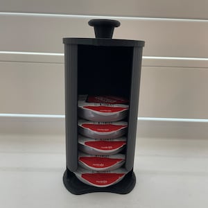 May include: A black plastic dispenser with a black knob on top holds six single-serve coffee pods. The pods are stacked vertically inside the dispenser.