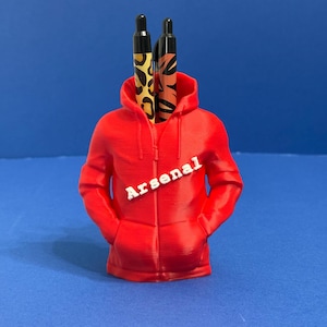 May include: A red hoodie-shaped pen holder with the word "Arsenal" printed on the front. Two pens with animal print designs are placed inside the hood. The background is a solid blue.