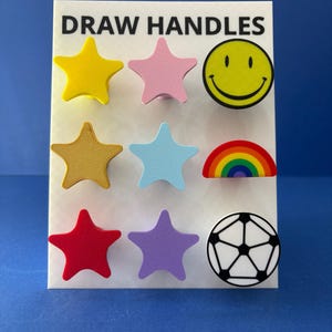 May include: A display card labelled DRAW HANDLES featuring nine decorative knobs in various shapes and colours. The set includes six star-shaped knobs in yellow, pink, gold, light blue, red, and purple, plus a yellow smiley face, a rainbow, and a football.