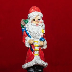 May include: A Christmas-themed figurine of Santa Claus atop a tree trunk with a small house. Santa wears a red coat, hat, and holds a sack. The tree trunk has a door, window, and is covered in snow.
