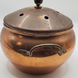 Peut inclure: Une marmite en cuivre avec un couvercle. La marmite a un corps arrondi avec une poignée de chaque côté. Le couvercle est d'une couleur cuivre plus foncée avec trois petits trous et un petit bouton sur le dessus. La marmite est probablement utilisée pour cuisiner ou servir des aliments.