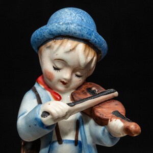 Puede incluir: Una figurita de porcelana de un niño que lleva un sombrero azul y ropa azul y blanca, tocando el violín. La figurita está de pie sobre una base marrón.