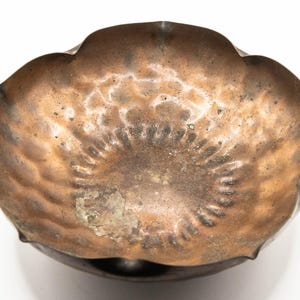 May include: A copper-toned bowl with a scalloped edge and a dark, textured exterior. The interior has a mottled, aged appearance with lighter and darker brown tones. The bowl's shape is reminiscent of a flower or a shell, and it sits on a dark base.