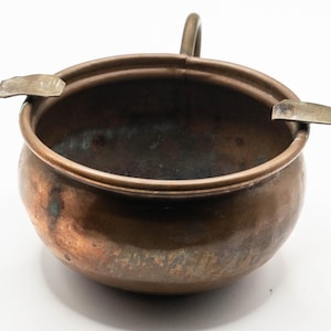 Peut inclure: Un cendrier vintage en cuivre avec une petite poignée et deux supports intégrés. Le cendrier a une forme de bol arrondie et une patine ternie, montrant des signes d'âge et d'utilisation. La couleur générale est un mélange de cuivre et de bronze.