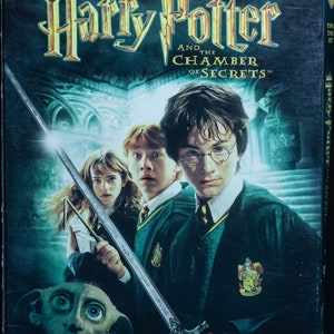 May include: DVD cover for the movie Harry Potter and the Chamber of Secrets. The cover features a dark green background with a silver sword and a close-up of Harry Potter, Ron Weasley, and Hermione Granger. The title of the movie is in gold lettering.