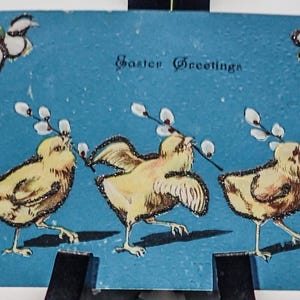 May include: Vintage Easter postcard with a blue background and three yellow chicks carrying pussy willow branches. The words "Easter Greetings" are written across the top. Decorative flowers and glitter accents frame the image.