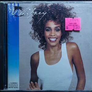 May include: A compact disc case with a photo of Whitney Houston on the front. The case is blue and white with the text "Whitney" in a white font. The price tag is pink and says "Sam's Jams Phone 547-SAMS $7.99".