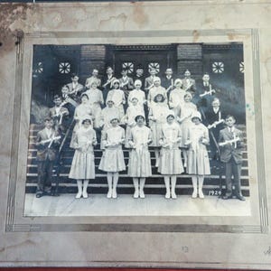 May include: A black and white photograph of a group of children, likely a graduating class, standing on a set of stairs in front of a building. The children are wearing formal attire and holding small bouquets of flowers. The photo is dated 1929.