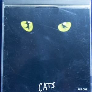 May include: A black CD case with a close-up of two yellow cat eyes and the word "CATS" in white text. The text "ACT ONE" is in the bottom right corner.