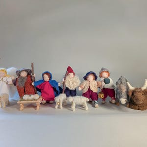 May include: A collection of handmade felted nativity figures, including an angel, shepherds, and the three wise men. The figures are dressed in colorful clothing and are accompanied by a lamb, a donkey, and an ox. The set is arranged on a white surface.