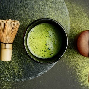 Könnte beinhalten: Eine schwarze Keramikschale gefüllt mit leuchtend grünem Matcha-Tee, begleitet von einem hellen Bambusbesen und einem langen, schmalen Bambuslöffel, der auf einem dunklen Holzdeckel liegt, alles bestäubt mit feinen grünen Matcha-Pulverpartikeln.