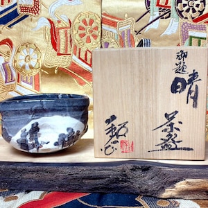May include: A ceramic tea bowl with a dark grey and white glaze featuring abstract dark markings, positioned next to a square wooden storage box. The box lid displays black Japanese calligraphy and a small red seal stamp on the front surface.