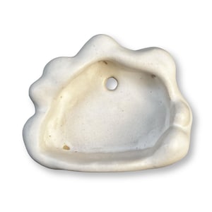 May include: Off-white ceramic decorative dish with a wavy, scalloped edge. The dish has a shallow basin and a small hole in the centre. The surface has subtle colour variations, suggesting a handmade or aged appearance.