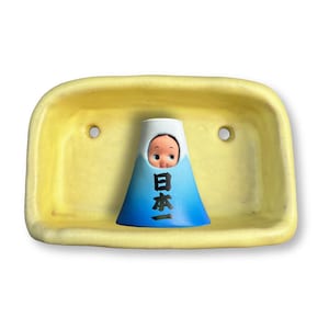 Puede incluir: Un jabonera de cerámica amarilla con dos agujeros, que sostiene una figura azul y blanca con forma del Monte Fuji. La figura tiene una cara de muñeca y texto japonés. Un artículo decorativo único.