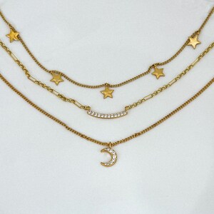 May include: A layered gold-toned necklace featuring three strands. The top strand has star charms, the middle has a curved bar with clear stones, and the bottom has a crescent moon pendant with clear stones. The necklace is on a white background.