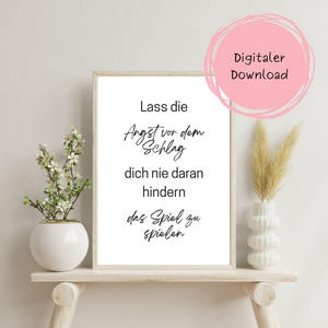 May include: A framed print with German text, displayed on a wooden shelf with decorative vases and plants. The text reads "Lass die Angst vor dem Schlag dich nie daran hindern das Spiel zu spielen." A pink circle with "Digitaler Download" is in the upper right.