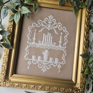 May include: A framed cross-stitch artwork featuring a white design on a tan background. The design includes candles, keys, and a decorative shelf. The gold frame has ornate detailing. Green foliage surrounds the frame, and a pair of scissors rests below.