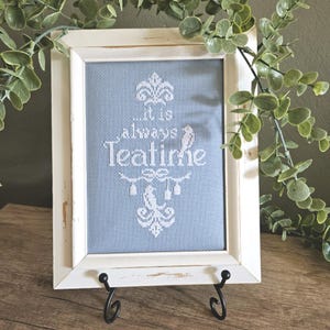May include: A framed cross-stitch piece featuring white embroidery on a light blue fabric background. The text reads "...it is always Teatime" and is surrounded by decorative white bird and floral motifs. The frame is white with a distressed finish.
