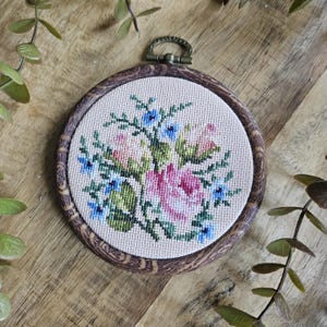 Op de afbeelding: Een ronde borduurring met een donkere houtnerfafwerking met daarin een kruissteekwerk op lichtbeige stof. Het ontwerp toont een centraal boeket van roze rozen omringd door kleine blauwe bloemetjes en groene bladeren.