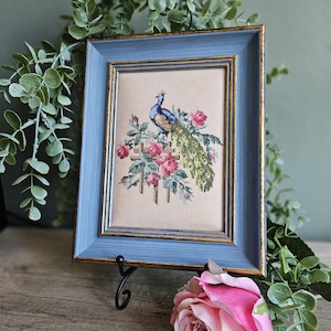 Op de afbeelding: Een ingelijst kruissteek borduurwerk met een blauwe en groene pauw op een houten hek, omringd door roze rozen. Het kunstwerk zit in een rechthoekige blauwe lijst met goudkleurige binnenranden en toont gedetailleerd naaldwerk.