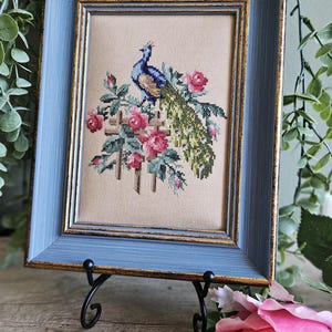 May include: A cross-stitch embroidery featuring a blue and green peacock perched on a wooden fence surrounded by pink roses. The artwork is displayed in a rectangular blue frame with gold inner trim, set on a black metal stand.