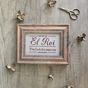 Op de afbeelding: Een ingelijst kruissteekwerk met de woorden "El Roi" en "The God who sees me" in bordeauxrode draad. De lijst is van rustiek hout. Decoratieve gedroogde bloemen en een kleine zilveren schaar zijn rond de lijst op een houten oppervlak geplaatst.