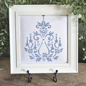 Op de afbeelding: Een vierkante witte lijst met een kruissteekborduurwerk op witte stof. Het ontwerp toont een symmetrisch blauw patroon met bloemmotieven, kaarsvormen en decoratieve krullen die in het midden van de stof zijn geplaatst.