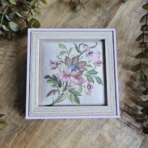 Op de afbeelding: Een vierkant kruissteek borduurwerk met een bloemmotief in roze, bruine, blauwe en groene draden op een lichte stoffen achtergrond, geplaatst in een lichtgekleurde houten lijst met een zachtroze buitenrand.