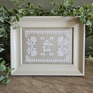 May include: A framed cross-stitch embroidery featuring white thread on a tan linen background. The design displays a central birdcage motif flanked by floral rose patterns and small bows, all enclosed within a decorative geometric border.