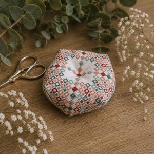 Op de afbeelding: Een rond, opgevuld speldenkussen met een geometrisch kruissteekpatroon in tinten rood, roze en groenblauw op een witte stoffen ondergrond. Het speldenkussen heeft een centrale inkeping en wordt vergezeld door een kleine zilverkleurige metalen schaar.