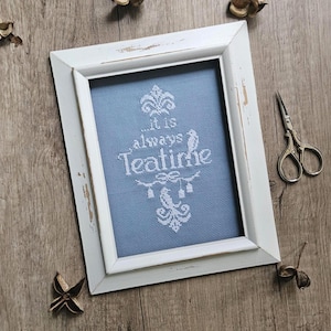 May include: A framed cross-stitch with the text "It is always Teatime" in white on a blue background. The white frame has a distressed finish. Dried seed pods and small scissors are arranged around the frame on a wooden surface.