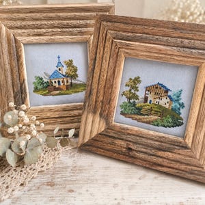 Op de afbeelding: Twee ingelijste kruissteekkunstwerken. Eén toont een kerk met een blauw dak en een boom, de andere een huis met een blauw dak en bomen. Beide bevinden zich in rustieke houten frames, met een landelijk thema.