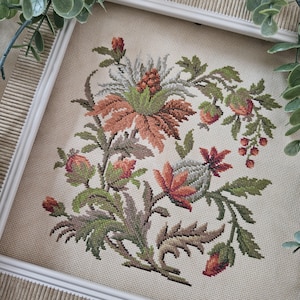 Op de afbeelding: Een kruissteek borduurwerk met een botanisch arrangement van gestileerde bloemen, knoppen en bladeren in tinten roestoranje, saliegroen en gedempt grijs, op een lichtbeige stoffen achtergrond in een witte lijst.