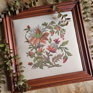 May include: A square cross-stitch embroidery featuring a botanical design with orange, red, and green flowers and leaves on a cream-colored fabric background, encased in a polished dark brown wooden frame with a beveled edge.