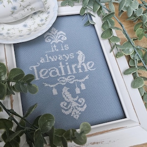 May include: A framed cross-stitch featuring the words "...it is always Teatime" in white thread on a blue fabric. The white frame has a distressed appearance. A teacup and saucer with floral patterns are visible in the top left.