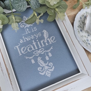 May include: A framed cross-stitch with the words "...it is always Teatime" in white on a blue fabric. The white frame has a distressed look. A floral teacup and saucer are visible in the background.