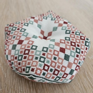 Op de afbeelding: Een handgemaakt, kruissteek kussen met een geometrisch patroon in tinten wit, rood, groen en roze. Het kussen heeft een afgeronde vorm en ligt op een licht houten oppervlak. De steken zijn gedetailleerd en precies.