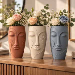Op de afbeelding: Drie cilindrische vazen in de vorm van menselijke gezichten met gesloten ogen en lippen. De vazen zijn terracotta, crème en leiblauw van kleur en bevatten elk een boeket rozen, gipskruid en eucalyptusbladeren op een houten oppervlak.