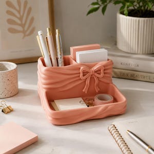 May include: A peach-colored ceramic desk organizer featuring a sculpted bow detail on the front. The organizer has a tall back section with two compartments for pens and sticky notes, and a lower front tray for small items like tape and clips.