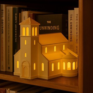 May include: A cream-colored church-shaped lamp with illuminated windows and a steeple. The lamp is placed on a wooden shelf among books. The warm light from the windows creates a cozy ambiance. The lamp is a decorative item.