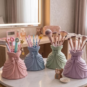 May include: Four pastel-colored decorative holders shaped like dresses, each filled with makeup brushes and pens. The holders are pink, blue, green, and purple, and are arranged on a white table. The holders have a bow detail.