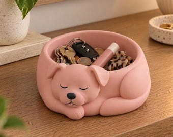 May include: A matte pink ceramic bowl shaped like a sleeping dog, featuring a smooth finish and a black nose. The interior of the bowl holds various small items including metal keys, a black car key fob, a pink lip balm, and a patterned hair clip.