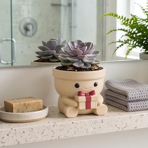 Op de afbeelding: Een lichtbeige keramische plantenpot in de vorm van een klein figuurtje dat een cadeau met een rood lint vasthoudt. De pot bevat een groene vetplant. Het figuurtje heeft eenvoudige zwarte ogen en een kleine glimlach, staand op een gespikkeld aanrecht.