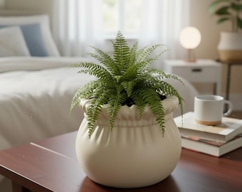 Op de afbeelding: Een groene varen in een crèmekleurige, ronde pot met een gerimpelde rand staat op een donkerbruine houten tafel. De plant is het middelpunt, met een slaapkamer op de achtergrond. De pot is crèmekleurig.