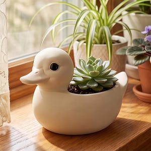May include: A matte off-white ceramic planter shaped like a duck with a smooth finish and dark eyes. The duck-shaped vessel contains a green succulent plant with thick, layered leaves planted in dark soil, positioned as a decorative container.