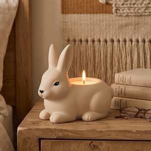 May include: A matte cream-coloured ceramic rabbit-shaped candle holder with a lit white candle inside its back. The rabbit has a smooth finish, pointed ears, and a small black nose. The candle flame is bright yellow and orange, casting a soft glow.