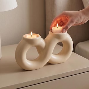 May include: Cream-colored ceramic candle holder with two circular openings, each holding a lit tea light candle. The holder has a smooth, curved design. A hand is placing a lit candle into the second opening. The holder is on a beige surface.