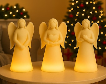 May include: Three illuminated angel figurines with outstretched wings, each with a different hand position. The angels are a soft yellow color and sit on a round table. A Christmas tree with lights and ornaments is in the background.
