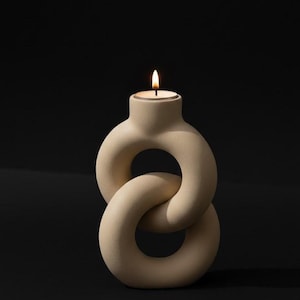 May include: A beige ceramic candle holder featuring two interlocking circular rings. The top ring includes a cylindrical opening that holds a small, lit tea light candle with a visible flame. The surface of the ceramic has a matte, textured finish.