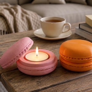 May include: A pink macaron-shaped candle holder with a lit tea light candle inside and a separate pink lid leaning against it, alongside a closed orange macaron-shaped container. Both items feature a textured, cookie-like exterior design.